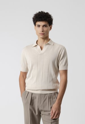 Joven con cabello rizado oscuro que lleva una camisa de punto beige de manga corta y pantalones plisados marrón claro, de pie con las manos en los bolsillos.