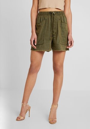 Frau trägt olivgrüne Shorts mit Kordelzug und großen Fronttaschen sowie hohe Sandalen in Nude, beiger bauchfreier Top sichtbar.