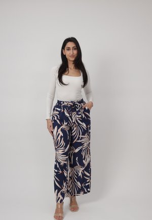 Kvinde stående, iført hvid langærmet top og marineblå bukser med brede ben og beige bladmotiv, poserer mod en ensfarvet lys baggrund.