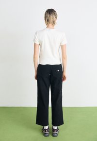 T-shirt bianco a maniche corte, pantaloni larghi neri con tasche posteriori e sneakers grigie con dettagli rosa, su uno sfondo neutro.