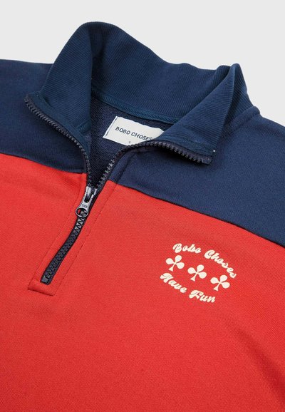 Half-zip sweatshirt with navy collar and red body, featuring "Bobo Choses Have Fun" text and three club suit symbols on the chest.