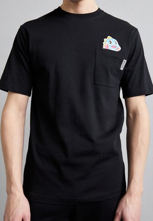 Persoon draagt een zwart t-shirt met een kleine kleurrijke cartoon dinosaurus die uit het borstzakje aan de linkerkant gluurt.