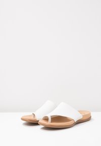 White open-toe sandals with a wide, curved upper and a tan rubber sole, featuring a textured braid detail on the side.