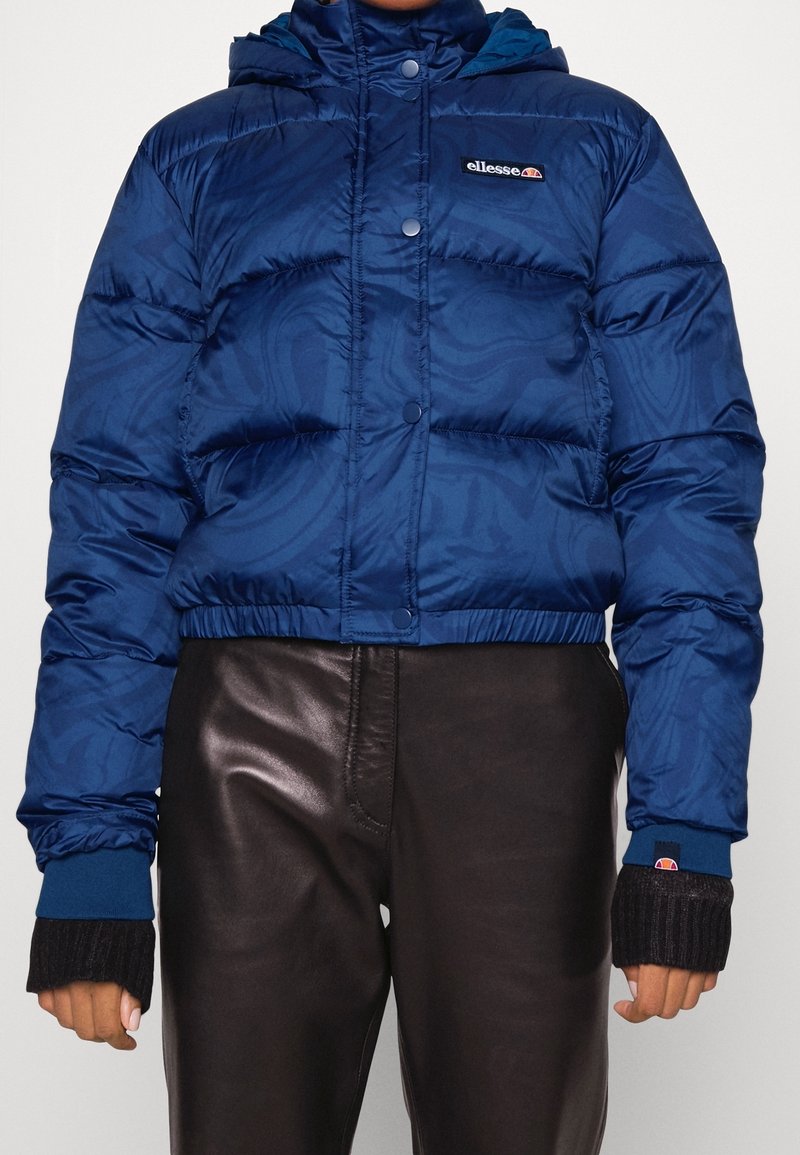 Niebieska kurtka puchowa z wzorzystą teksturą, skrócona długość, zapięcie na guziki i kaptur. Posiada ściągacze przy mankietach oraz logo na klatce piersiowej.
