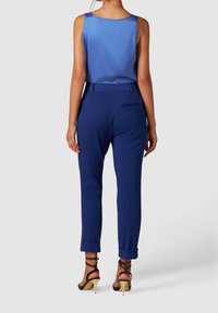 Femme portant un chemisier bleu sans manches, un pantalon taille haute bleu marine avec revers, et des sandales à talons hauts noires à lanières, debout de dos.