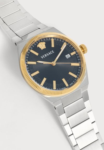 Montre-bracelet Versace avec bracelet en métal argenté, cadran noir, index et aiguilles dorés, lunette dorée, logo Méduse et guichet date à 3 heures.