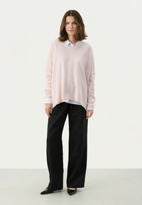 Maglione oversize rosa chiaro in tessuto morbido, indossato sopra una camicia bianca con colletto. Abbinato a pantaloni neri a gamba larga e tacchi neri affilati.