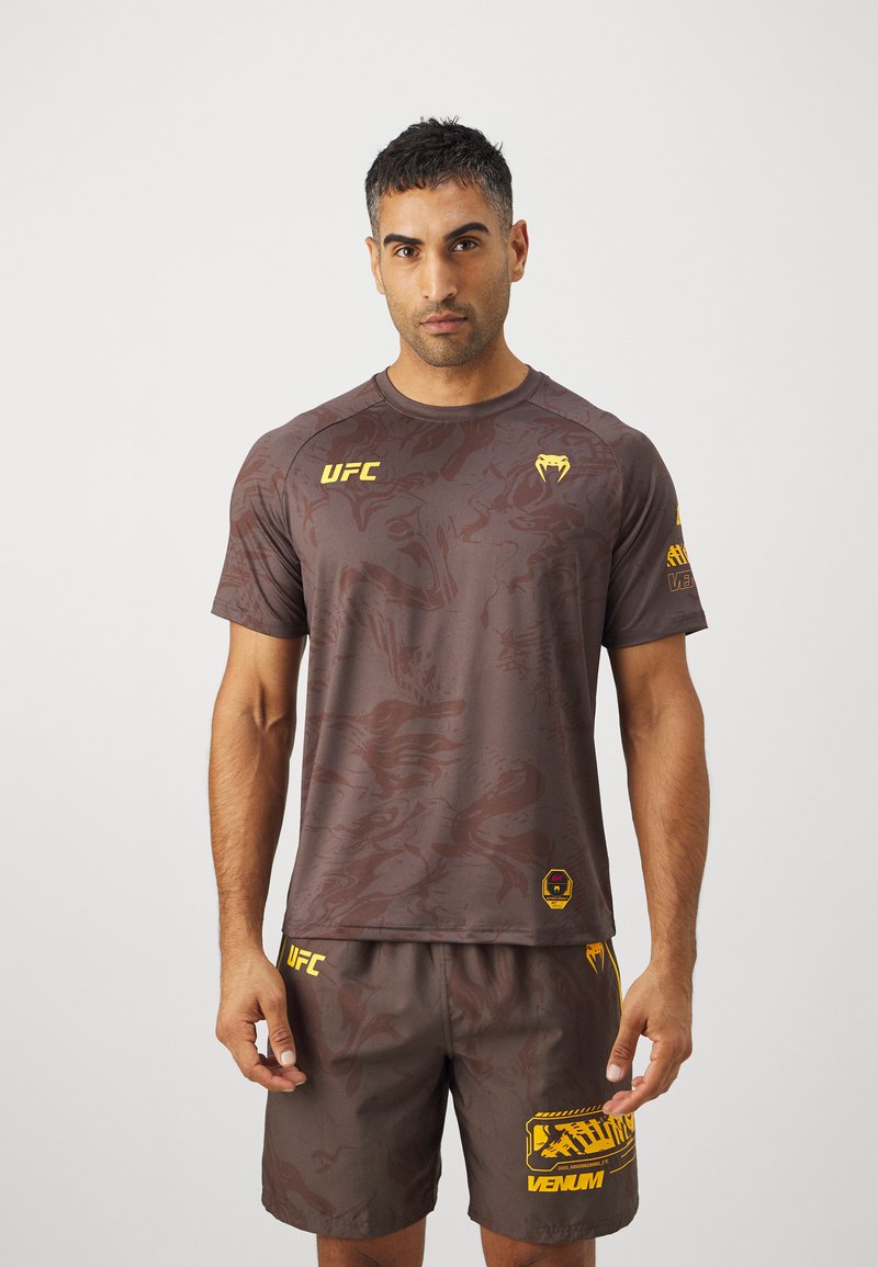 Camiseta deportiva marrón con un patrón sutil, logotipo amarillo de UFC en el pecho, y pantalones cortos a juego con acentos amarillos y un diseño gráfico.