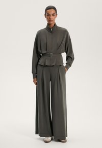 Oliwkowy dres z zamkiem błyskawicznym i paskiem w talii oraz spodniami o szerokich nogawkach. Gładki materiał, luźny krój oraz delikatne fałdy.