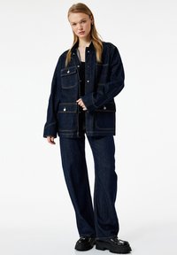 Ciemnoniebieska kurtka dżinsowa z czterema przednimi kieszeniami, srebrnymi zatrzaskami i kontrastującym żółtym przeszyciem, zestawiona z luźnymi ciemnoniebieskimi spodniami dżinsowymi.