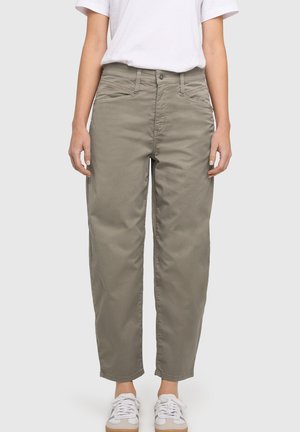 Persoon draagt een olijfgroene rechte broek met hoge taille, witte sneakers en een wit T-shirt met korte mouwen tegen een effen achtergrond.