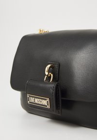 Melnas ādas rokassomi taisnstūra formā, ar zelta detaļām un priekšējo atloku. Izceļas ar zelta "LOVE MOSCHINO" birku un nelielu spriegojuma detaļu.