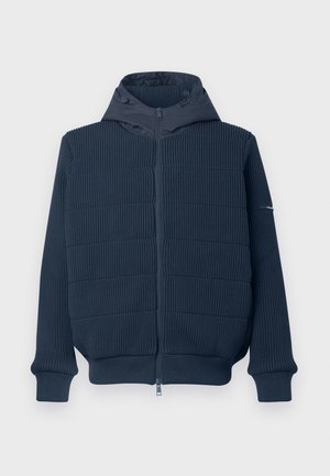 Laivastonsininen vetoketjuhuppari, jossa on uritettu pinta, sileä huppu ja joustavat hihansuut. Edessä on vetoketju ja yksityiskohta yhdessä hihassa.