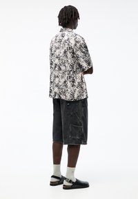 Camicetta a maniche corte con motivo in nero e beige, abbinata a pantaloni corti grigi scuri con tasche. Sandali neri con cinghie alla caviglia completano il look.