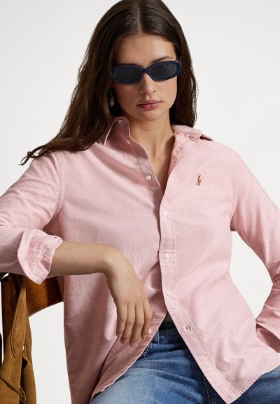 Svetloružová košeľa na gombíky z jemnej látky s malým vyšívaným logom. Oblečená s tmavými slnečnými okuliarmi, sediaca pri drevenej stoličke.