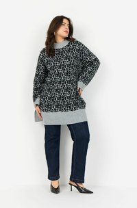 Frau trägt einen langen Pulli mit Leopardenmuster, grauen Bündchen, dunkelblauen Jeans und schwarzen Absatzmules und posiert mit einer Hand auf der Hüfte.