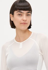 Hvid mesh langærmet top med V-udskæring, der har raglanærmer og en minimal mærke detalje foran. Let og struktureret design.