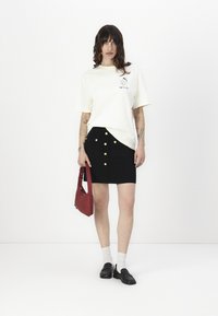White t-shirt with graphic print, black skirt with front buttons, black loafers, white socks, and red handbag. Plain background.