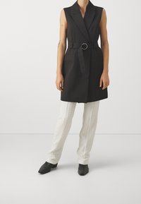 Černá bezrukávová vesta s opaskem se zaoblenou sponou, kombinovaná s texturovanými bílými kalhotami a černými botami s špi�čatou špičkou.