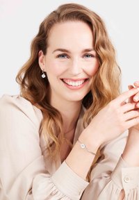 Zilveren armband met cirkelvormige edelsteendetail, gedragen om een pols, gecombineerd met hangende oorbellen met een soortgelijk ontwerp. Lichte stof blouse.