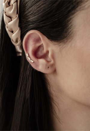 Silberner Ohrclip mit herzförmigen Akzenten, vor glatter Haut, mit einem geflochtenen Haarband im Hintergrund.