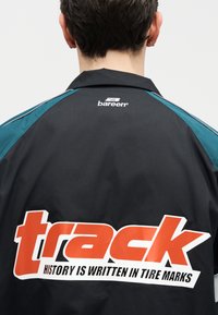 Άτομο που φοράει μαύρο μπουφάν με πράσινες λεπτομέρειες, με μεγάλο πορτοκαλί κείμενο "track" και το σύνθημα "Η ΙΣΤΟΡΙΑ ΓΡΑΦΕΤΑΙ ΣΕ ΣΗΜΑΔΙΑ ΕΛΑΣΤΙΚΩΝ" στην πλάτη.