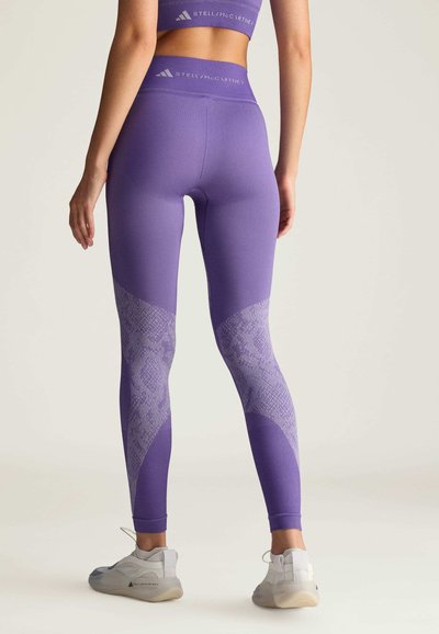 Purple leggings featuring a textured snakeskin pattern at the lower legs and a high waistband. Paired with light grey athletic shoes.