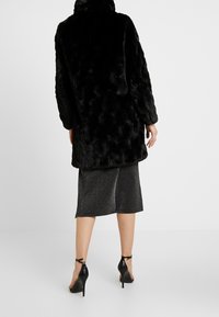 Sort faux fur frakke med lange ærmer, høj krave og en tekstureret overflade, parret med en metallisk grå nederdel og sorte højhælede sko.