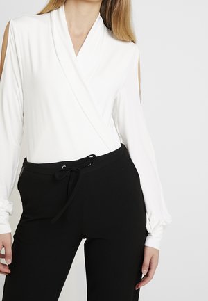 Frau trägt ein weißes Wickel-Oberteil mit langen Ärmeln und Schulterausschnitten sowie schwarze Hose mit Kordelzug vor einfarbigem Hintergrund.