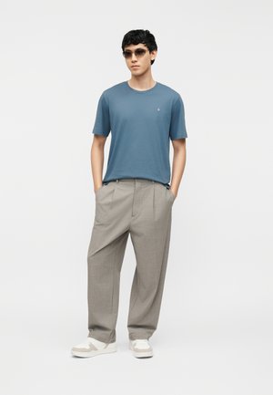 Nuori mies sininen t-paita, beige leveälahkeiset housut, valkoiset lenkkarit ja ruskeat aurinkolasit, seisoo käsivarsissa taskuissa.