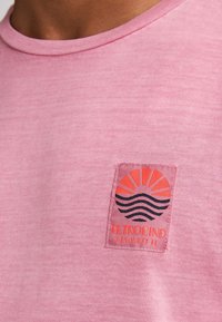 Rosa T-Shirt mit kurzen Ärmeln aus weichem Stoff, versehen mit einem aufgenähten Patch mit einem Sonnen- und Wellenmotiv sowie dem Text "PETROLIND KEY WEST FL".