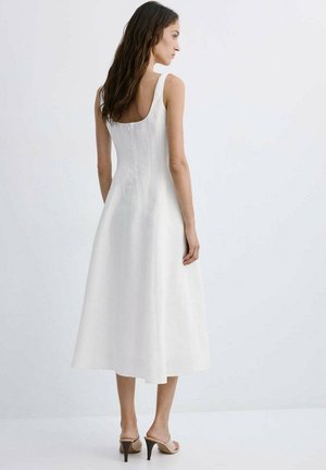 Mujer con el pelo largo y oscuro que lleva un vestido midi blanco sin mangas y sandalias beige de tacón, posando frente a un fondo claro y liso.