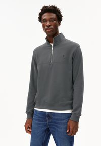 Sudadera gris con cremallera y cuello alto, con un logo sutil en el pecho. Combinada con jeans azules, mostrando un atuendo casual.