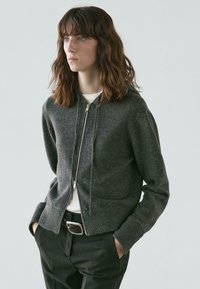 Dark gray zip-up hoodie made of soft material, featuring a kangaroo pocket and ribbed cuffs. Worn over a white shirt and black trousers.