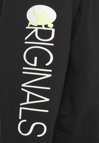 Svart sweatshirt med vit text "ORIGINALS" på ärmen och en ljusgrön grafik av en person. Mjukt tyg med ribbade muddar.