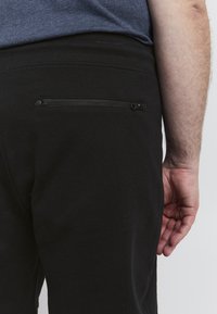 Pantalons de jogging noirs avec une texture lisse, dotés d'une poche zippée inclinée à l'arrière. La taille est élastique, offrant un ajustement parfait.