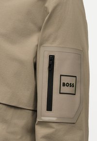 Μανίκι μπεζ μπουφάν με κάθετη μαύρη τσέπη με φερμουάρ και ορθογώνιο πατς με την ένδειξη «BOSS» στον άνω βραχίονα.