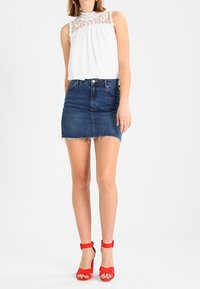 Witte blouse met kantdetails op de schouders, gecombineerd met een blauwe denim mini-jurk met een rafelige zoom, en rode sandalen met hoge hakken en banden.