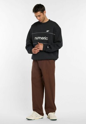 Mann trägt schwarzen "numeric"-Sweatshirt, braune Hose und weiße Turnschuhe, steht mit leicht gesenktem Kopf und verschränkten Händen.