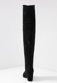 Mustat murskaava polven ylle ulottuvat korkokengät, joissa on lohkarepohja ja kaareva yläreuna, seisomassa valkoisella pinnalla yksinkertaista taustaa vasten.