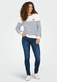 Pasiaste bluza z białymi i granatowymi paskami, z graficznym wzorem w postaci czerwonej wargi, zestawiona z niebieskimi obcisłymi dżinsami i białymi trampkami. Teksturowany materiał i swobodny krój.