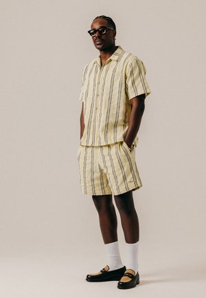 Homme portant une chemise à manches courtes rayée jaune assortie et un short, des lunettes de soleil, des chaussettes blanches et des mocassins, debout les mains dans les poches.