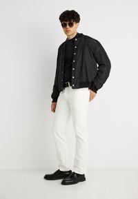 Νέος άνδρας με μαύρο μπουφάν τύπου bomber, μαύρο πουκάμισο, άσπρο παντελόνι, μαύρη ζώνη, μαύρα μοκασίνια και γυαλιά ηλίου, στέκεται μπροστά σε απλό λευκό φόντο.