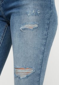 Μπλε τζιν παντελόνι με στενή εφαρμογή, με φθαρμένες περιοχές και σκισίματα στον μηρό. Η υφή είναι λεία με ξεθωριασμένο πλύσιμο.