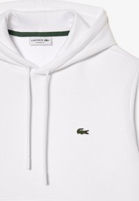 Sweat à capuche sans manches blanc avec cordons, arborant un logo crocodile vert Lacoste sur la poitrine et une étiquette intérieure indiquant "Lacoste Classic Fit".