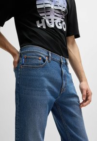 Blå denimjeans med rak skärning, fem fickor och orange sömnad. I kombination med en svart T-shirt med grafisk design.