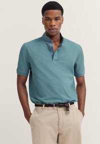 Polohemd aus teal Baumwolle mit kurzen Ärmeln, verfügt über einen kontrastierenden blauen Kragen, eine Knopfleiste mit zwei Knöpfen und ein besticktes Logo auf der Brust.