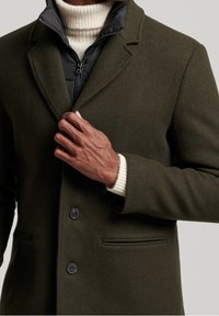 Manteau en laine vert olive avec une coupe ajustée, revers crantés, et deux boutons noirs. Superposé à une veste intérieure noire à fermeture éclair.