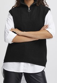 Černý žebrovaný bezrukávový svetr s vysokým límcem a stříbrným zipem, nošený přes bílou košili. Měkká textura a uvolněný střih.