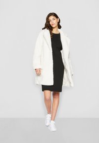 Cappotto in pelliccia sintetica bianca con una finitura testurizzata, indossato sopra un vestito nero e sneaker bianche. Il cappotto ha una vestibilità ampia e rever larghi.